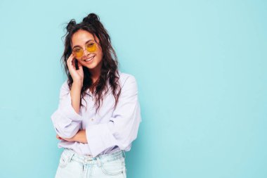 Moda kıyafetleri içinde gülümseyen güzel bir kadın. Stüdyoda mavi duvarın yanında poz veren kaygısız seksi kadın. Pozitif esmer model eğleniyor ve deliriyor. Neşeli ve mutlu güneş gözlükleriyle
