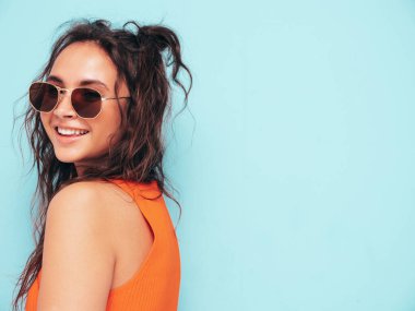 Moda kıyafetleri içinde gülümseyen güzel bir kadın. Stüdyoda mavi duvarın yanında poz veren kaygısız seksi kadın. Pozitif esmer model eğleniyor ve deliriyor. Neşeli ve mutlu güneş gözlükleriyle