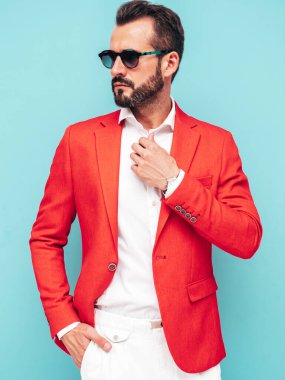 Yakışıklı, kendine güvenen, stil sahibi, lamberseksüel modelin portresi. Zarif kırmızı takım elbise giymiş modern bir adam. Moda erkeği stüdyoda mavi duvarın yanında güneş gözlüklü poz veriyor.
