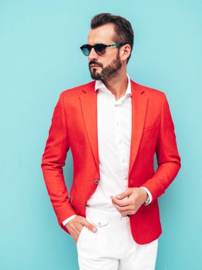 Yakışıklı, kendine güvenen, stil sahibi, lamberseksüel modelin portresi. Zarif kırmızı takım elbise giymiş modern bir adam. Moda erkeği stüdyoda mavi duvarın yanında güneş gözlüklü poz veriyor.