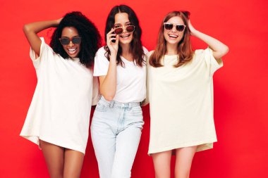 Moda kıyafetleri içinde üç güzel, gülümseyen uluslararası hippi kadın. Stüdyoda kırmızı duvarın yanında poz veren kaygısız kadınlar. Pozitif modeller güneş gözlükleriyle eğleniyor. Arkadaşlık kavramı