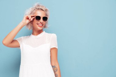 Son moda beyaz elbiseler içinde gülümseyen güzel bir kadının portresi. Stüdyoda mavi duvarın yanında poz veren kaygısız sarışın kadın. Pozitif model içeride eğleniyor. Neşeli ve mutlu güneş gözlükleriyle