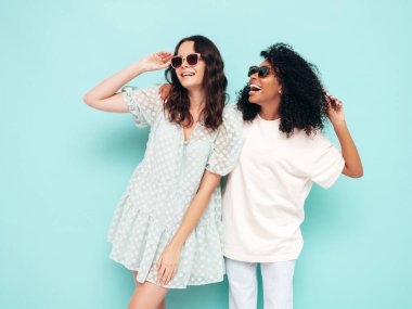Moda kıyafetleri içinde iki güzel, gülümseyen uluslararası hippi kadın. Stüdyoda mavi duvarın yanında poz veren kaygısız seksi kadınlar. Pozitif modeller eğleniyor. Arkadaşlık kavramı