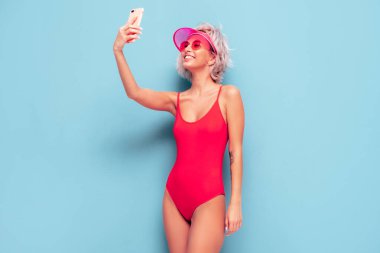 Yazın mayolu, gülümseyen sarışın manken kırmızı mayo ve şeffaf şapka giyiyor. Seksi kaygısız kadın eğleniyor ve deliriyor. Stüdyoda mavi duvarın yanında poz veren bir kadın. Selfie çekiyorum. 