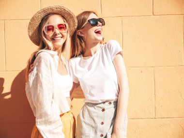 Son moda yaz kıyafetleri içinde iki güzel gülümseyen yeniyetme kadın. Sokakta şapkalı kayla poz veren kaygısız kadınlar. Pozitif saf modeller gün batımında eğleniyorlar, sarılıyorlar. 