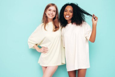 Moda kıyafetleri içinde iki güzel, gülümseyen uluslararası hippi kadın. Stüdyoda mavi duvarın yanında poz veren kaygısız seksi kadınlar. Pozitif modeller eğleniyor. Arkadaşlık kavramı. Güneş gözlükleriyle