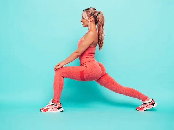 Fitness smiling woman in pink sports clothing. Young beautiful model with perfect body.Female posing near blue wall in studio.Cheerful and happy. Stretching out before training. Doing lunges