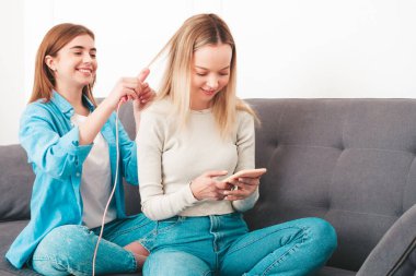 İki güzel, güzel, gülümseyen kadın. Lüks bir dairede ya da otel odasında, kanepede oturan kaygısız mankenler. Makyaj yapmak ve güzellik tedavisi yapmak. Saç düzleştirici kullanarak.