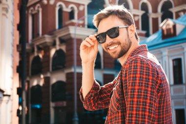 Yakışıklı, şık, havalı, havalı, lamberseksüel model. Kırmızı kareli gömlek giymiş modern bir adam. Moda erkeği güneş gözlükleriyle gökdelenin yanında poz veriyor. Gün batımında dışarıda. 
