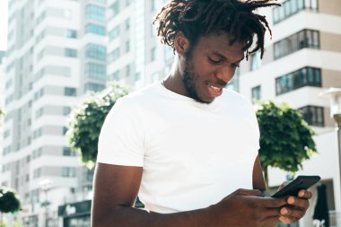 Yakışıklı hippi mankeni. Beyaz yaz tişörtü giymiş traşsız Afrikalı bir adam. Rasta saçlı, moda bir erkek. Sokakta poz vermek. Akıllı telefon ekranına bakmak, cep telefonu uygulamaları kullanmak.