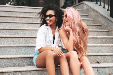 Son moda yaz kıyafetleri içinde iki güzel, gülümseyen kadın. Sokakta poz veren kaygısız, çok ırklı kadınlar. Güneş gözlüklü, pozitif modeller. Neşeli ve mutlu. Merdivenlerde oturuyor. 