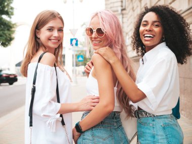 Son moda yaz kıyafetleri içinde gülümseyen üç genç bayan. Sokakta poz veren kaygısız, çok ırklı kadınlar. Güneş gözlükleriyle eğlenen pozitif modeller. Neşeli ve mutlu