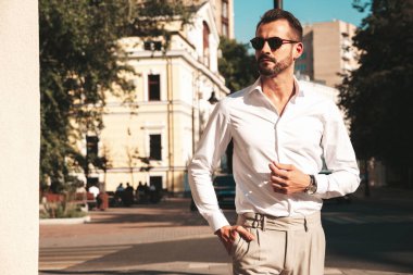 Yakışıklı, kendine güvenen, stil sahibi, lamberseksüel modelin portresi. Beyaz gömlek ve pantolon giymiş modern bir adam. Moda erkeği gün batımında Avrupa 'da sokak arkasındaymış gibi poz veriyor. Güneş gözlükleriyle