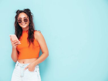 Son moda turuncu tişörtlü ve kot pantolonlu güzel, gülümseyen bir kadın. Stüdyoda mavi duvarın yanında poz veren kaygısız seksi kadın. Akıllı telefon ekranına bakıyorum. Cep telefonu uygulamaları kullanan bir model