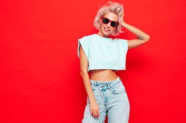 Moda kıyafetleri içinde gülümseyen güzel bir kadın. Stüdyoda kırmızı duvarın yanında poz veren kaygısız seksi kadın. Pozitif sarışın model eğleniyor ve deliriyor. Neşeli ve mutlu. Güneş gözlükleriyle