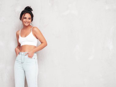 Moda kıyafetleri içinde gülümseyen güzel bir kadın. Stüdyoda gri duvarın yanında poz veren kaygısız seksi kadın. Pozitif esmer model eğleniyor ve deliriyor. Neşeli ve mutlu