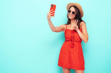 Son moda kırmızı elbisesi içinde gülümseyen güzel bir kadın. Stüdyoda mavi duvarın yanında poz veren kaygısız seksi kadın. Pozitif esmer model eğleniyor. Güneş gözlüğü ve şapkasıyla. Barış işareti gösteriyor. Selfie çekiyor.