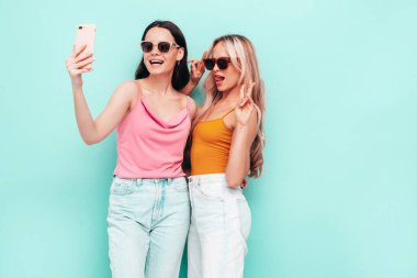 Moda kıyafetleri içinde gülümseyen iki güzel esmer hippi kadının portresi. Mavi duvarın yanında poz veren kaygısız seksi kadınlar. Pozitif modeller eğleniyor. Neşeli ve mutlu. selfie çekmek 