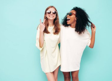 Moda kıyafetleri içinde iki güzel, gülümseyen uluslararası hippi kadın. Stüdyoda mavi duvarın yanında poz veren kaygısız seksi kadınlar. Pozitif modeller eğleniyor. Arkadaşlık kavramı