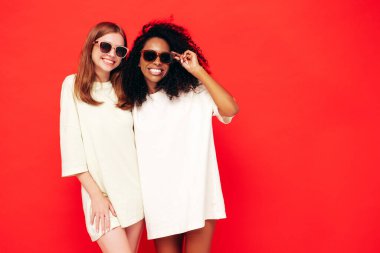 Moda kıyafetleri içinde iki güzel, gülümseyen uluslararası hippi kadın. Stüdyoda kırmızı duvarın yanında poz veren kaygısız seksi kadınlar. Pozitif modeller eğleniyor. Arkadaşlık kavramı. Güneş gözlükleriyle