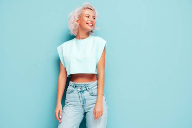 Son moda beyaz elbiseler içinde gülümseyen güzel bir kadının portresi. Stüdyoda mavi duvarın yanında poz veren kaygısız sarışın kadın. Pozitif model içeride eğleniyor. Neşeli ve mutlu