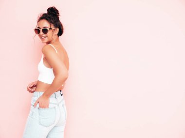 Moda kıyafetleri içinde gülümseyen güzel bir kadın. Stüdyoda pembe duvarın yanında poz veren kaygısız seksi kadın. Pozitif esmer model eğleniyor ve deliriyor. Neşeli ve mutlu 