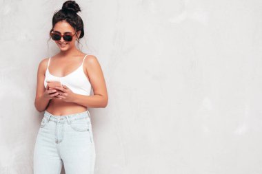 Moda kıyafetleri içinde gülümseyen güzel bir kadın. Stüdyoda gri duvarın yanında poz veren kaygısız seksi kadın. Pozitif esmer model eğleniyor ve deliriyor. Akıllı telefon uygulamaları kullanarak. Telefon bekletiliyor