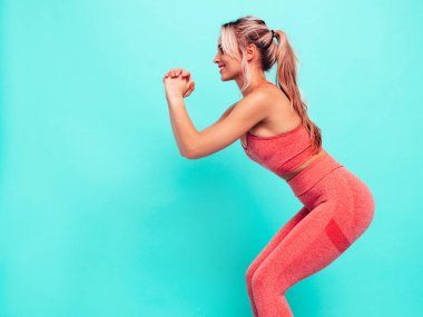 Pembe spor elbiseli, gülümseyen kadın. Mükemmel vücutlu genç, güzel bir manken. Stüdyoda mavi duvarın yanında poz veren bir kadın. Neşeli ve mutlu. Eğitimden önce esneme hareketleri. Çömeliyor.  