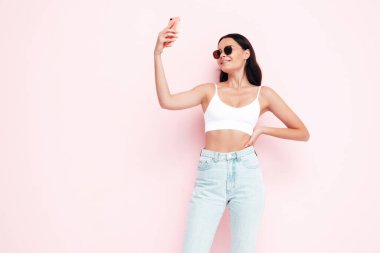Moda kıyafetleri içinde gülümseyen güzel bir kadının yakın plan portresi. Stüdyoda pembe duvarın yanında poz veren kaygısız seksi kadın. Pozitif model içeride eğleniyor. Selfie çekiyoruz. Güneş gözlükleriyle