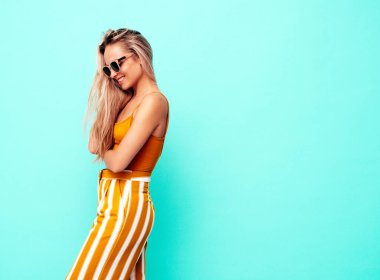 Son moda sarı elbiseli, gülümseyen, güzel sarışın kadın. Stüdyoda mavi duvarın yanında poz veren kaygısız seksi kadın. Pozitif model içeride eğleniyor. Neşeli ve mutlu. Güneş gözlükleriyle