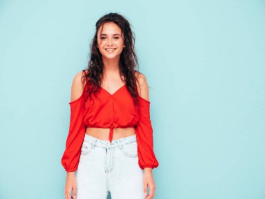 Genç, güzel, güler yüzlü, son moda kırmızı bluzlu ve kot elbiseli bir kadın. Stüdyoda mavi duvarın yanında poz veren kaygısız seksi kadın. Pozitif esmer model eğleniyor. Neşeli ve mutlu