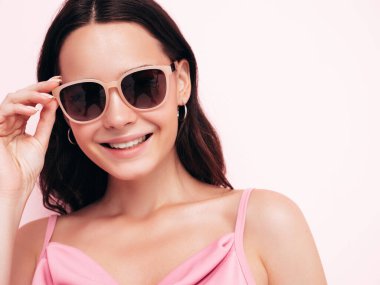 Moda kıyafetleri içinde gülümseyen güzel bir kadının yakın plan portresi. Stüdyoda pembe duvarın yanında poz veren kaygısız seksi kadın. Pozitif model içeride eğleniyor. Güneş gözlüğünü çıkarıyorum.