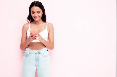 Moda kıyafetleri içinde gülümseyen güzel bir kadın. Stüdyoda pembe duvarın yanında poz veren kaygısız seksi kadın. Pozitif esmer model cep telefonu ekranına bakıyor. Akıllı telefonu tutmak ve uygulamaları kullanmak