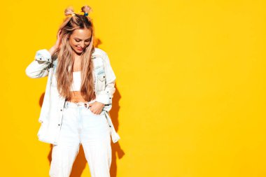 Moda kıyafetleri içinde gülümseyen sarışın güzel bir kadının portresi. Stüdyoda sarı duvarın yanında poz veren kaygısız seksi kadın. Pozitif model içeride eğleniyor. Neşeli ve mutlu