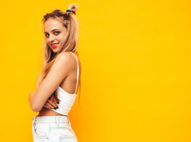 Moda kıyafetleri içinde gülümseyen sarışın güzel bir kadının portresi. Stüdyoda sarı duvarın yanında poz veren kaygısız seksi kadın. Pozitif model içeride eğleniyor. Neşeli ve mutlu