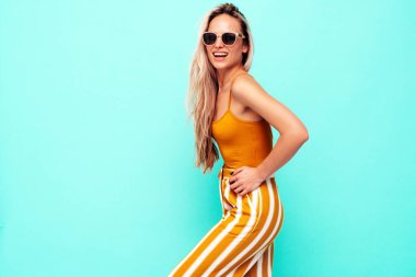 Son moda sarı elbiseli, gülümseyen, güzel sarışın kadın. Stüdyoda mavi duvarın yanında poz veren kaygısız seksi kadın. Pozitif model içeride eğleniyor. Neşeli ve mutlu. Güneş gözlükleriyle