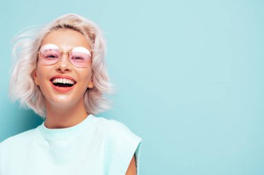 Son moda beyaz elbiseler içinde gülümseyen güzel bir kadının portresi. Stüdyoda mavi duvarın yanında poz veren kaygısız sarışın kadın. Pozitif model içeride eğleniyor. Neşeli ve mutlu güneş gözlükleriyle