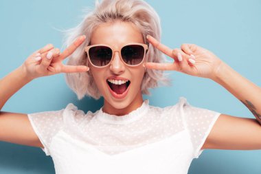 Moda kıyafetleri içinde gülümseyen güzel bir kadın. Stüdyoda mavi duvarın yanında poz veren kaygısız seksi kadın. Pozitif sarışın model güneş gözlükleriyle eğleniyor. Çığlıklar ve bağırışlar