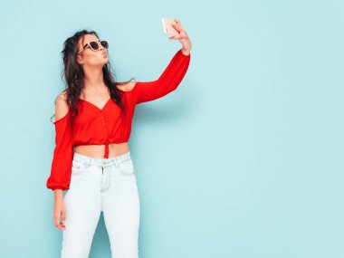 Genç, güzel, güler yüzlü, son moda kırmızı bluzlu ve kot elbiseli bir kadın. Stüdyoda mavi duvarın yanında poz veren kaygısız seksi kadın. Pozitif esmer model eğleniyor. Güneş gözlüklü selfie çekiyoruz.