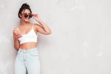 Moda kıyafetleri içinde gülümseyen güzel bir kadın. Stüdyoda gri duvarın yanında poz veren kaygısız seksi kadın. Pozitif esmer model eğleniyor ve deliriyor. Akıllı telefon uygulamaları kullanarak. Telefon bekletiliyor