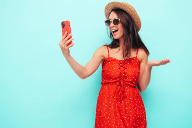 Son moda kırmızı elbisesi içinde gülümseyen güzel bir kadın. Stüdyoda mavi duvarın yanında poz veren kaygısız seksi kadın. Pozitif esmer model eğleniyor. Güneş gözlüğü ve şapkasıyla. Selfie çekiyor.