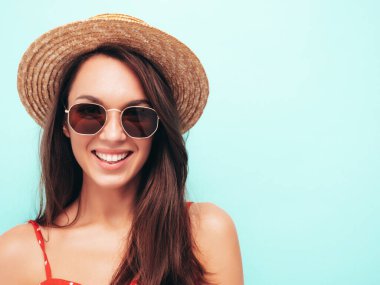 Moda bir yaz elbisesi içinde gülümseyen genç bir kadının yakın plan portresi. Stüdyoda mavi duvarın yanında poz veren kaygısız bir kadın. Kapalı alanda eğlenen olumlu bir model. Güneş gözlüğü ve şapkayla neşeli ve mutlu.