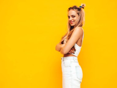 Moda kıyafetleri içinde gülümseyen sarışın güzel bir kadının portresi. Stüdyoda sarı duvarın yanında poz veren kaygısız seksi kadın. Pozitif model içeride eğleniyor. Neşeli ve mutlu