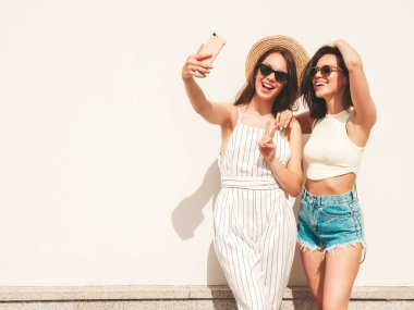 Son moda yaz kıyafetleri içinde iki güzel, gülümseyen hippi kadın. Sokakta beyaz şapkalı, kaygısız kadınlar poz veriyor. Pozitif saf modeller gün batımında eğleniyorlar, selfie çekiyorlar.