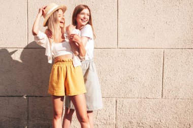 Son moda yaz kıyafetleri içinde iki güzel, gülümseyen hippi kadın. Sokakta beyaz duvarın yanında poz veren kaygısız kadınlar. Pozitif saf modeller gün batımında eğleniyorlar, sarılıyorlar ve deliriyorlar.