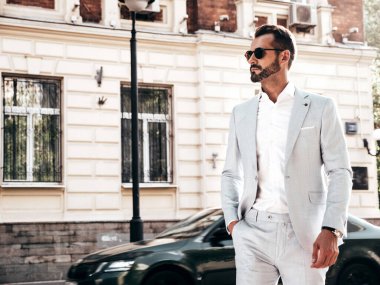 Yakışıklı, kendine güvenen, stil sahibi, lamberseksüel modelin portresi. Zarif beyaz takım elbise giymiş modern bir adam. Moda erkeği gün batımında Avrupa şehrinin arka planında poz veriyor. Güneş gözlükleriyle