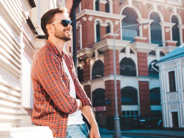 Yakışıklı, şık, havalı, havalı, lamberseksüel model. Kırmızı kareli gömlek giymiş modern bir adam. Moda erkeği güneş gözlükleriyle gökdelenin yanında poz veriyor. Gün batımında dışarıda. 