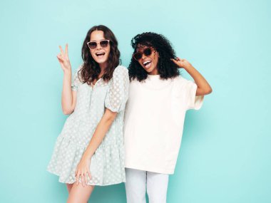 Moda kıyafetleri içinde iki güzel, gülümseyen uluslararası hippi kadın. Stüdyoda mavi duvarın yanında poz veren kaygısız seksi kadınlar. Pozitif modeller eğleniyor. Arkadaşlık kavramı