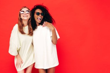 Moda kıyafetleri içinde iki güzel, gülümseyen uluslararası hippi kadın. Stüdyoda kırmızı duvarın yanında poz veren kaygısız seksi kadınlar. Pozitif modeller eğleniyor. Arkadaşlık kavramı. Güneş gözlükleriyle