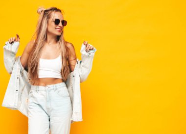 Moda kıyafetleri içinde gülümseyen sarışın güzel bir kadının portresi. Stüdyoda sarı duvarın yanında poz veren kaygısız seksi kadın. Pozitif model içeride eğleniyor. Neşeli ve mutlu güneş gözlükleriyle 